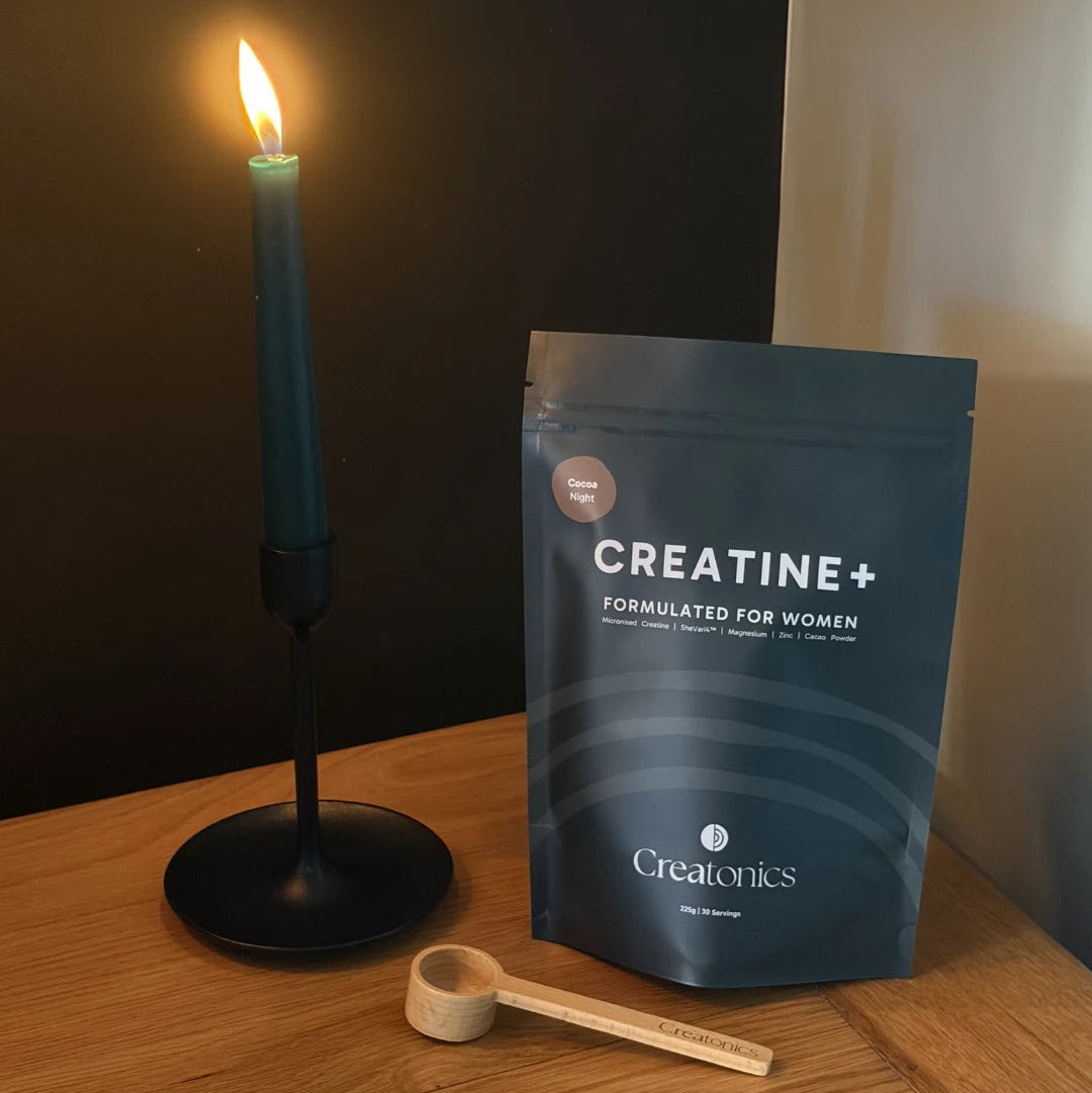 A stand-up pouch of Creatine+ formulated for women by Creatonics sits on a wooden table next to a lit teal candle and a small wooden scoop.