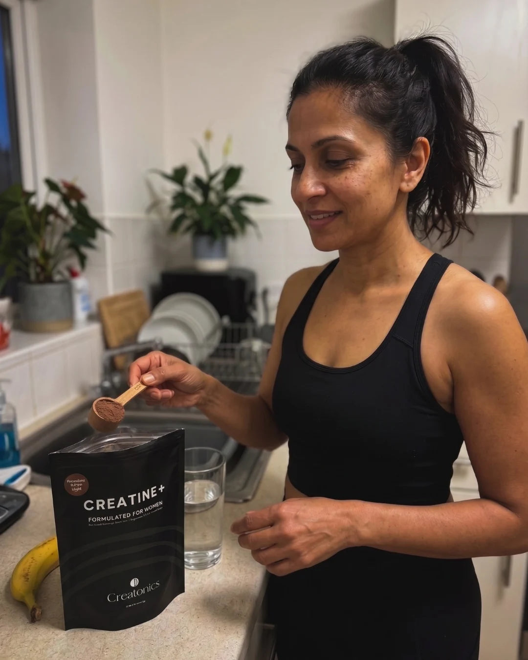 A woman in athletic wear scooping chocolate creatine powder from a black pouch of Creatonics Creatine+ Formulated for Women Cocoa Night in a kitchen setting.