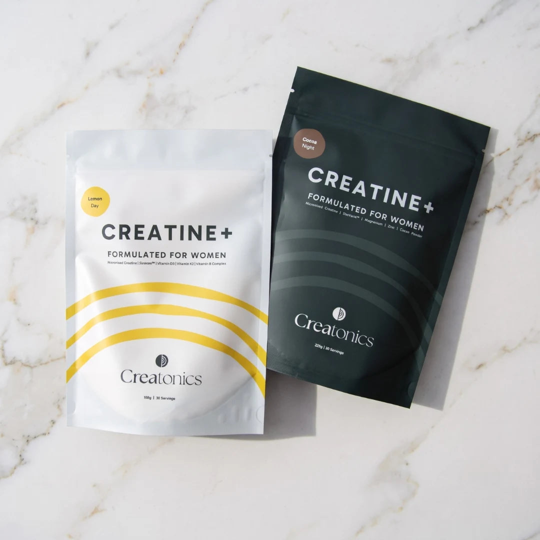 Two pouches of Creatonics Creatine+ Formulated for Women, the white Lemon Day pouch and the dark Cocoa Night pouch, displayed side-by-side on a light marble countertop