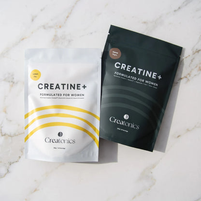 Two pouches of Creatonics Creatine+ Formulated for Women, the white Lemon Day pouch and the dark Cocoa Night pouch, displayed side-by-side on a light marble countertop