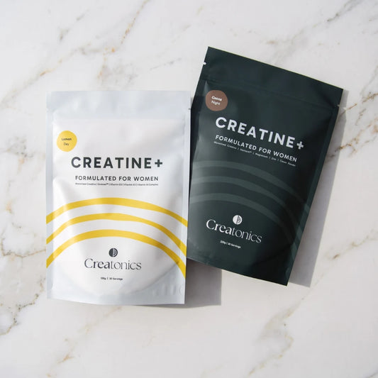 Two pouches of Creatonics Creatine+ Formulated for Women, the white Lemon Day pouch and the dark Cocoa Night pouch, displayed side-by-side on a light marble countertop