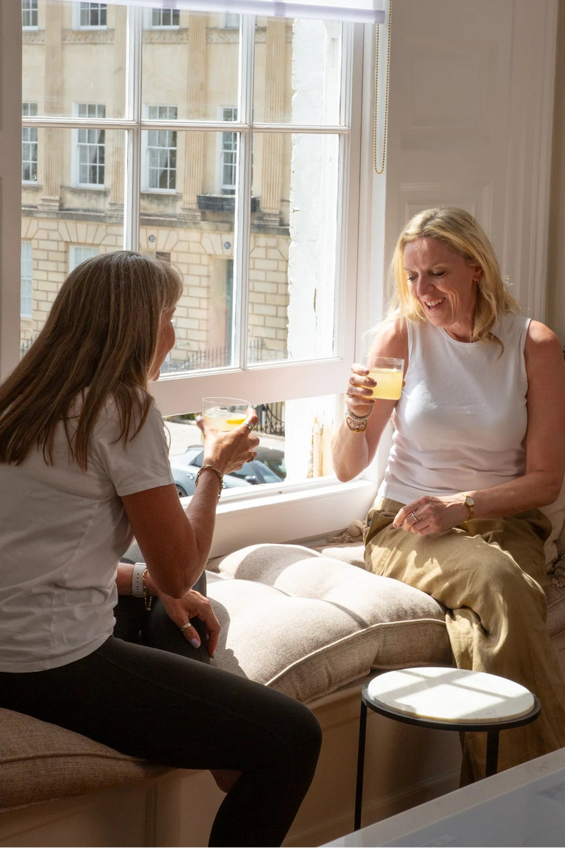 Two women sitting on a cushioned window seat indoors, each holding a Creatonics lemon drink. The room has warm neutral walls, a large bright window, and a small round side table between them.