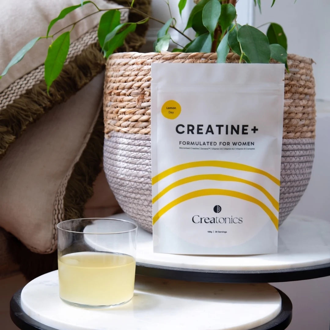 Creatonics Creatine+ Formulated for Women Lemon Day supplement pouch displayed next to a glass of mixed lemon creatine drink on a marble table with a potted plant.