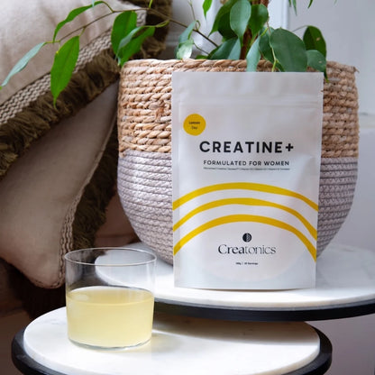 Creatonics Creatine+ Formulated for Women Lemon Day supplement pouch displayed next to a glass of mixed lemon creatine drink on a marble table with a potted plant.